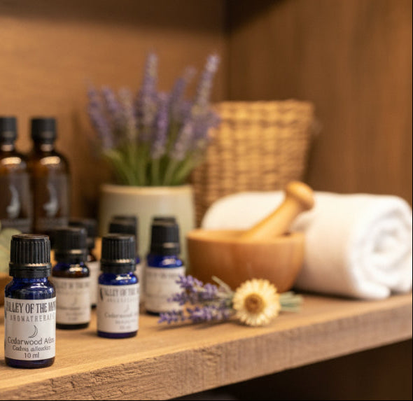 Collection of valley of the moon aromatherapy cedarwood atlas essential oil bottles, a mortar and pestle, and lavender flowers on a wooden shelf.