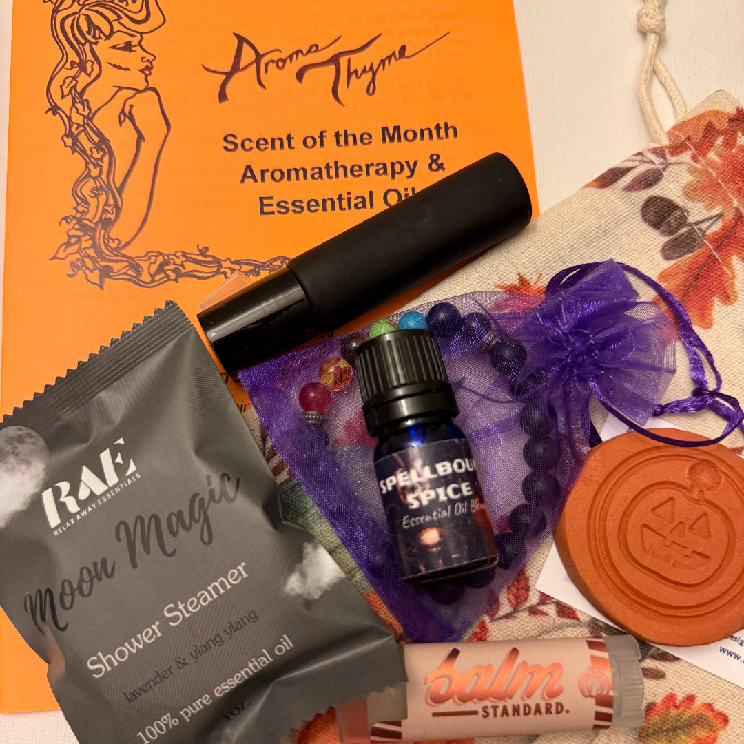 A curated autumn-themed aromatherapy and essential oil gift set arranged on a white surface. The set includes an orange Aroma Thyme 'Scent of the Month' card, a black roller  bottle, a gray RAE 'Moon Magic' shower steamer packet, a small blue 'Spellbound Spice' essential oil bottle, a terracotta pumpkin diffuser, a purple chakra beaded bracelet in a sheer purple bag, and a pumpkin spice  Balm Standard lip balm.